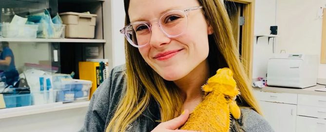 A smiling veterinarian with glasses holds a yellow bearded dragon lizard in a room filled with shelves and equipment.