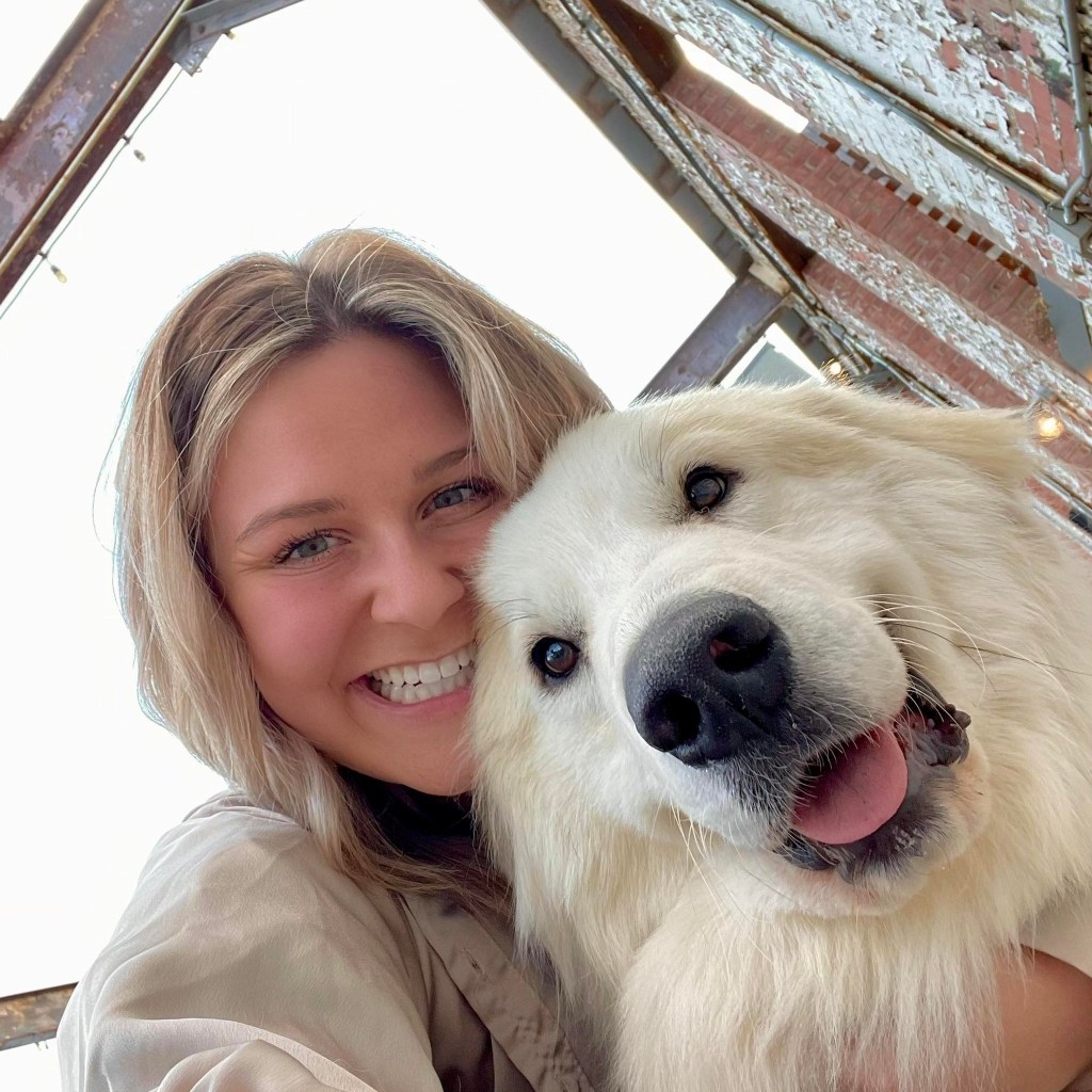 A blonde-haired person smiles while hugging a large, fluffy white dog near a vet clinic. They enjoy a cheerful moment outside together, with the dog looking content and its tongue slightly out.
