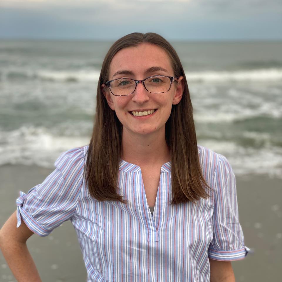 A veterinarian with glasses and long hair stands smiling on a beach, the ocean waves in the background. Wearing a striped shirt, their hands rest confidently on their hips, embodying the calm assurance of a trusted vet enjoying a well-deserved seaside break.