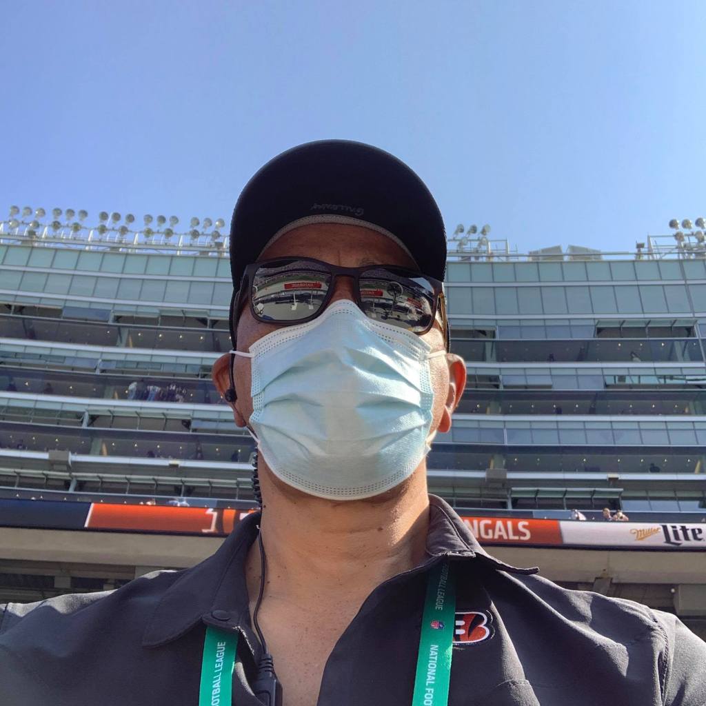 A person wearing a mask, sunglasses, and a cap stands outside the stadium. Dressed in a dark shirt with an event lanyard, the veterinarian seems ready for action under the clear blue sky as the multi-tiered arena looms in the background.