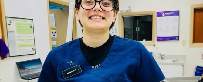 A veterinarian wearing glasses and a blue scrub top stands smiling in a veterinary clinic. The environment includes medical equipment, a laptop, and informational posters on the walls.