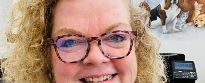 A person with curly blonde hair and glasses, likely a veterinarian, smiles at the camera. In the background, a mural depicts various animals, including a dog and a cat.