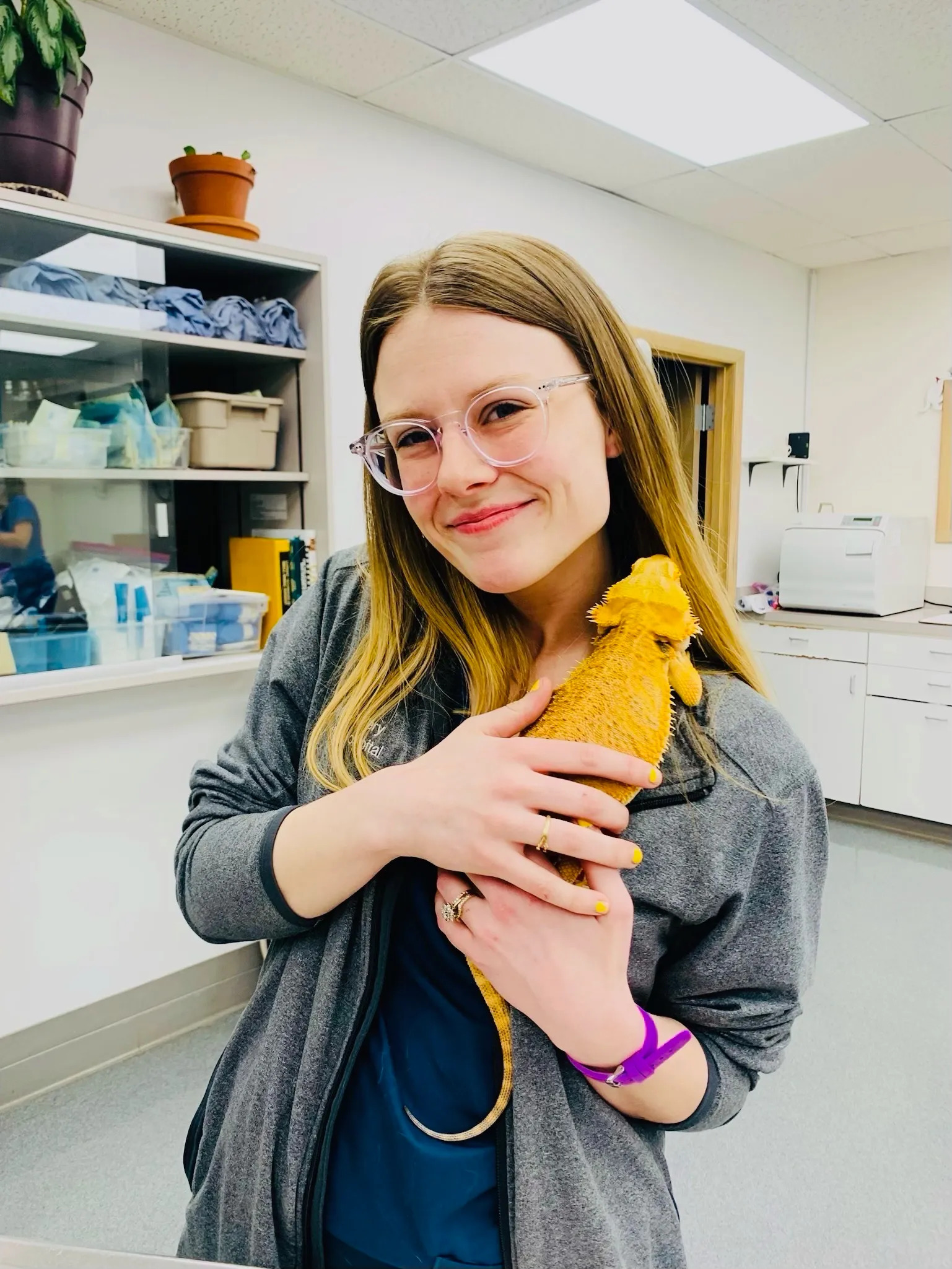 A woman with glasses and long hair smiles while holding an orange bearded dragon in a veterinary clinic. The veterinarian, wearing a gray jacket, stands against a backdrop of shelves filled with supplies and medical equipment.