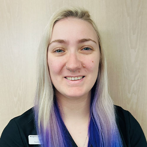 A person with long, straight, blonde hair fading to purple at the ends, wearing a black top and a nose ring, smiles at the camera in front of a light wood background.