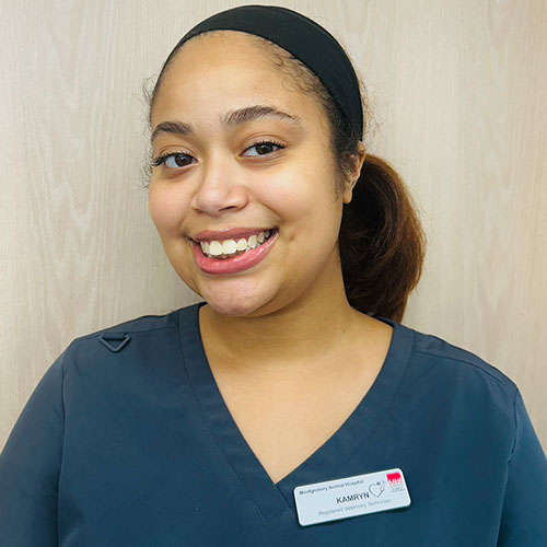 A woman with long brown hair tied back, wearing a dark gray scrub top and a black headband, smiles at the camera. She has a name badge clipped to her scrubs and stands in front of a light wood background.